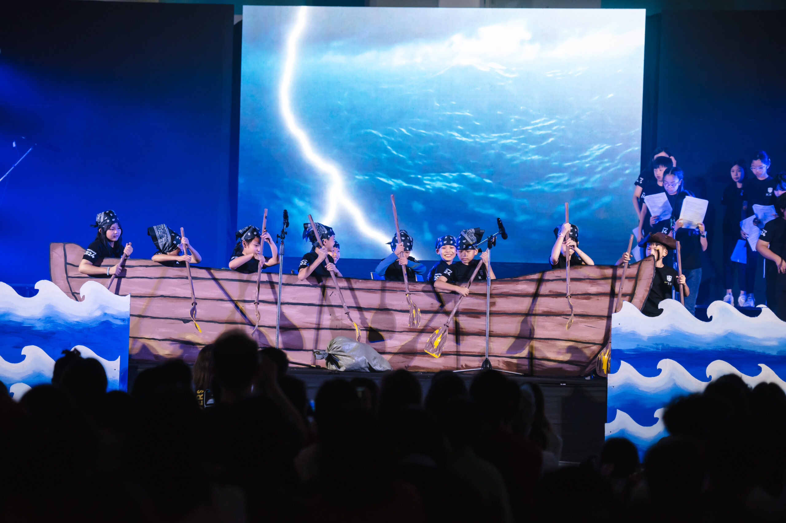Student performance reenacting a stormy sea scene with a boat prop, oars, and waves.