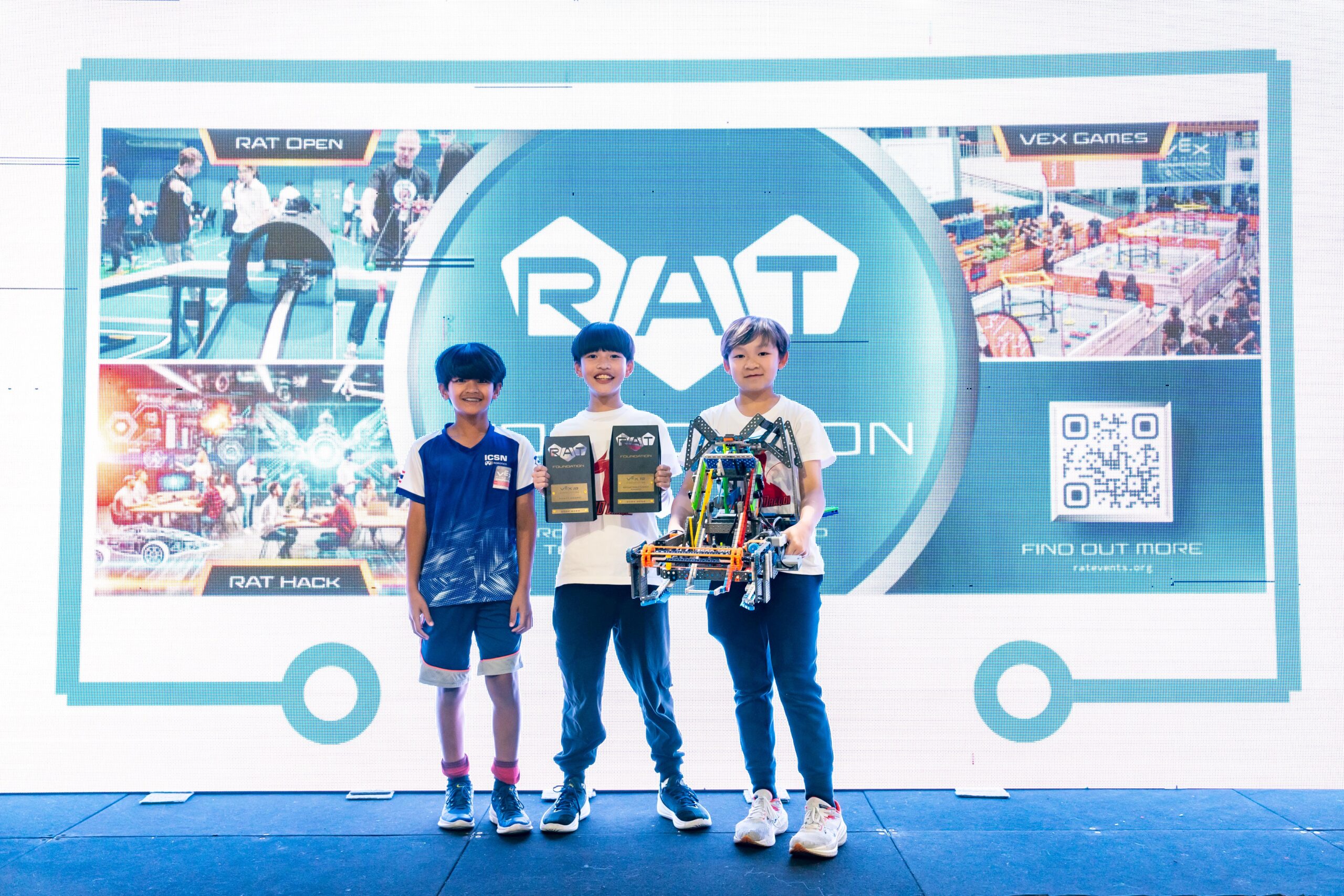 Three boys smiling while carrying the trophies and their robot during the awarding ceremony on stage.