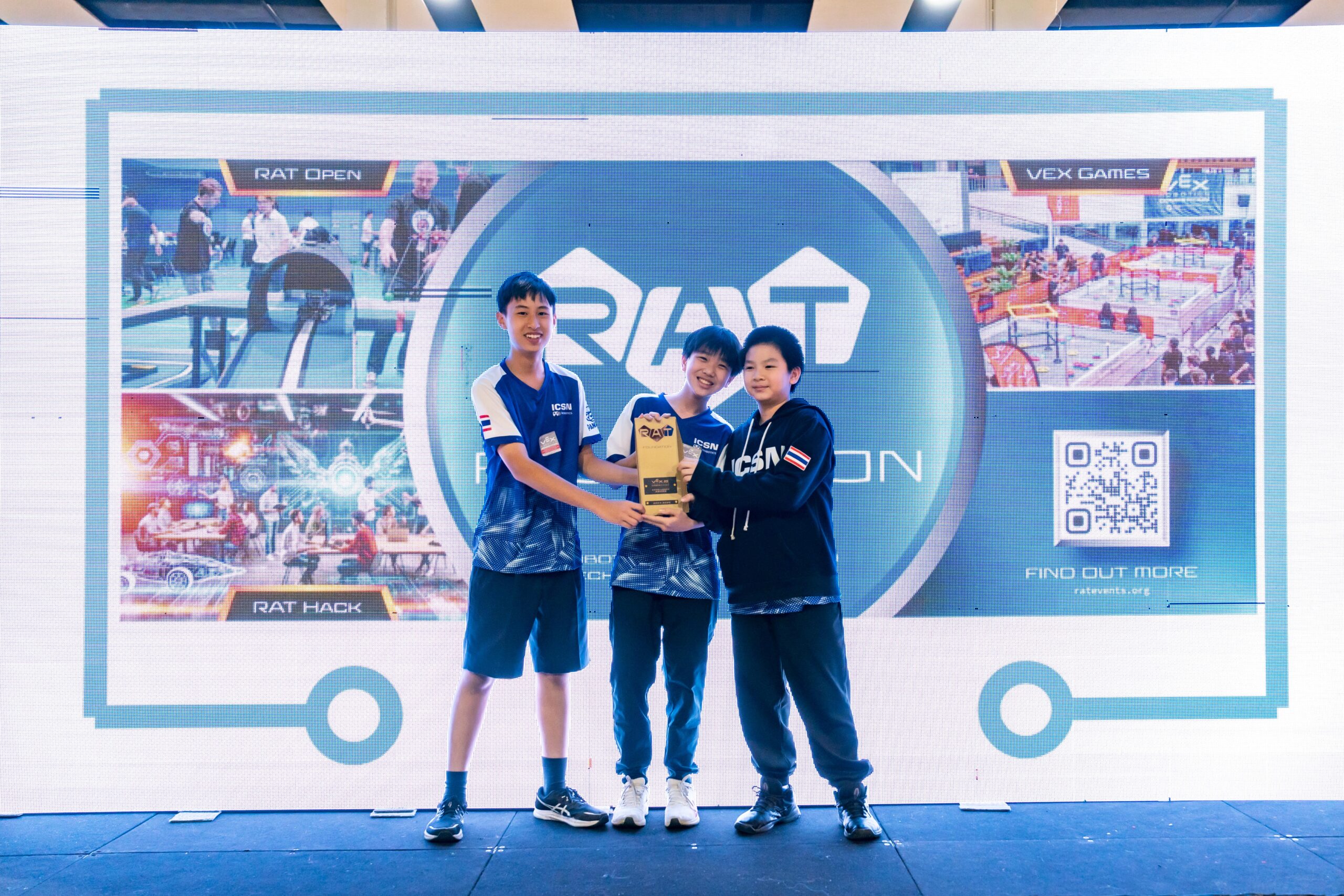 Three boys smiling, holding a gold trophy for winning an award at a robotics competition.