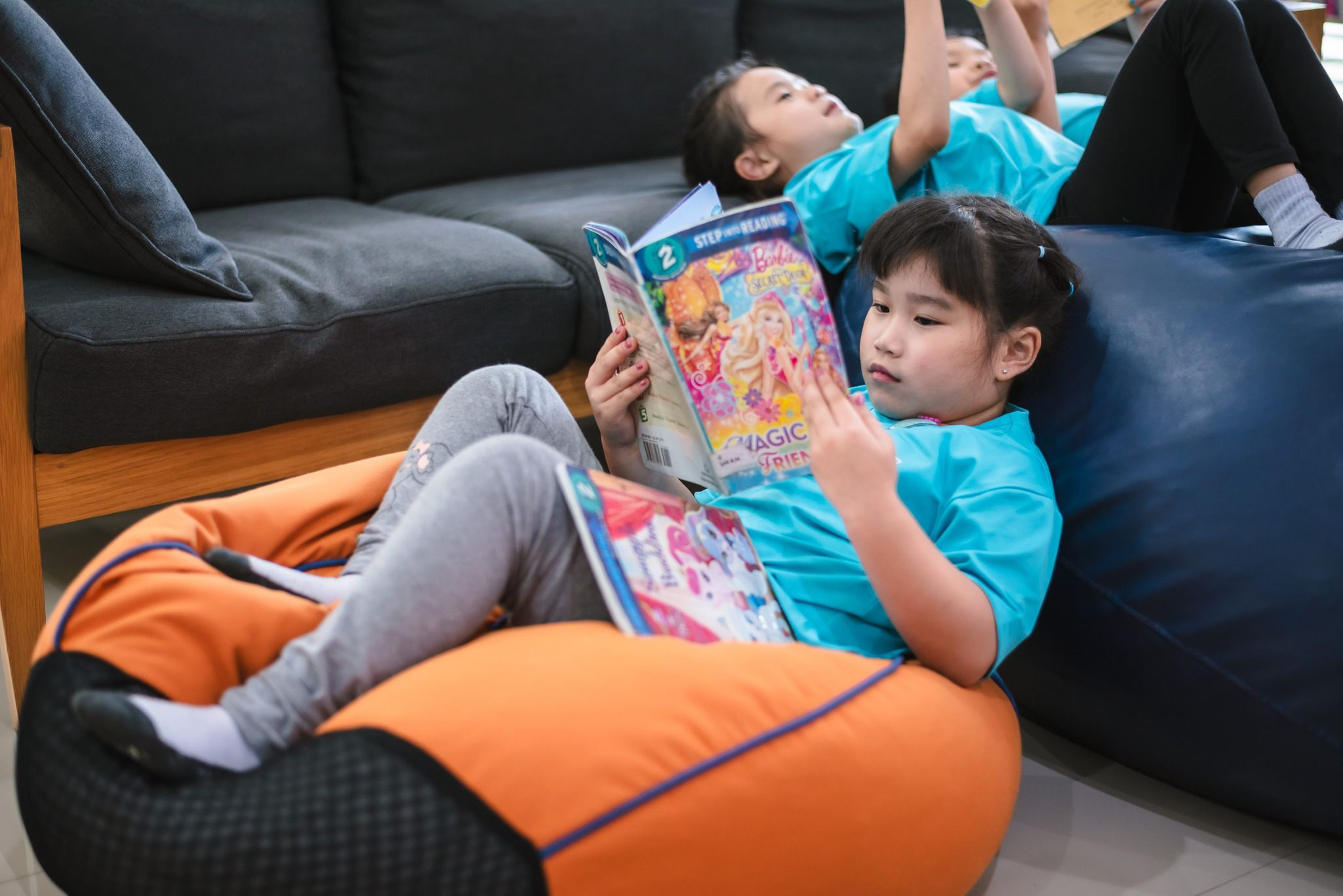 A girl laying on an orange bean bag while reading a book in the library with her classmates