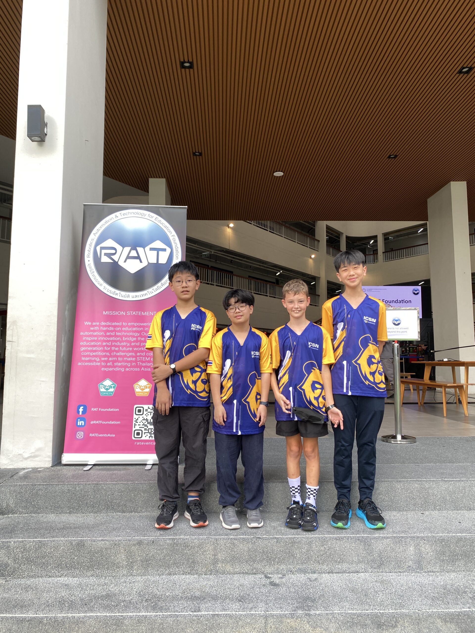 A group of four young boys in matching blue and yellow 'ICSN' team jerseys pose for a photo in a modern, multi-level building atrium.