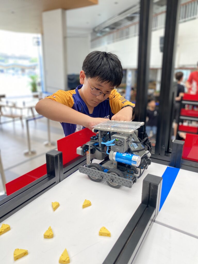 A focused young boy in an 'ICSN' team jersey and glasses bends over a competition field, using both hands to make adjustments to a gray and blue robot. Yellow, cheese-wedge-shaped game pieces are scattered on the white surface.