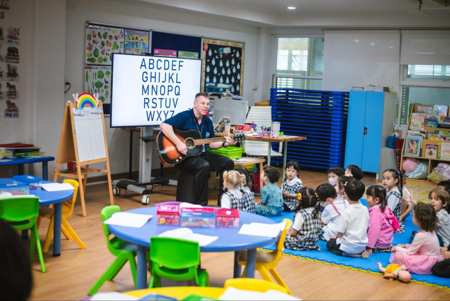 A kindergarten teacher teaching the alphabet song while playing his guitar in a colorful classroom