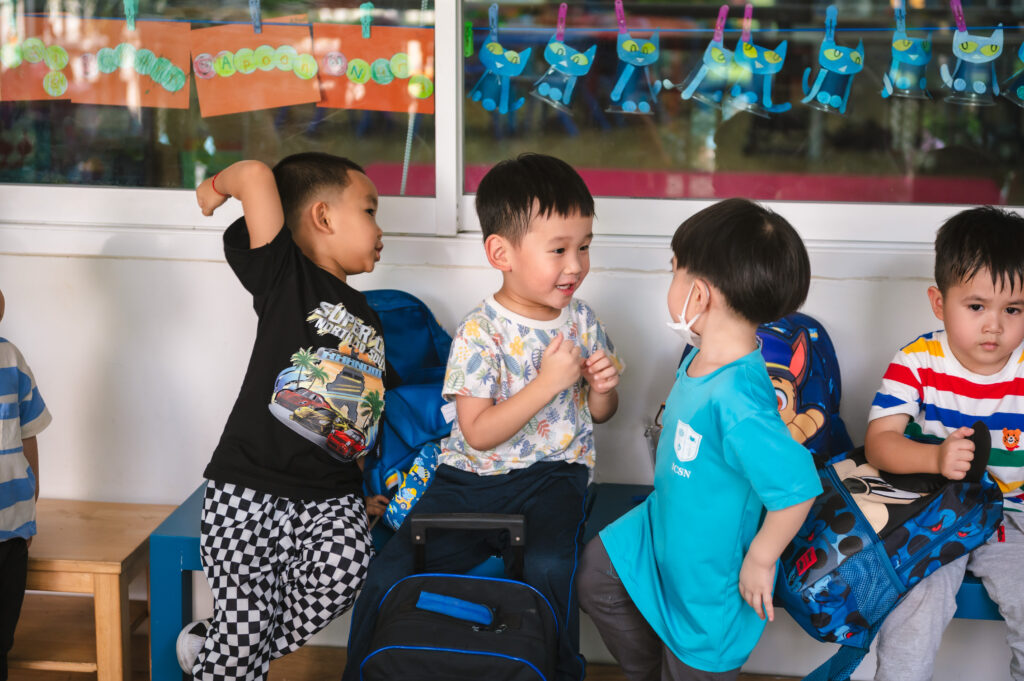Four young students at ICSN sit together on a blue bench, with two boys in the center engaged in an animated conversation and social interaction.