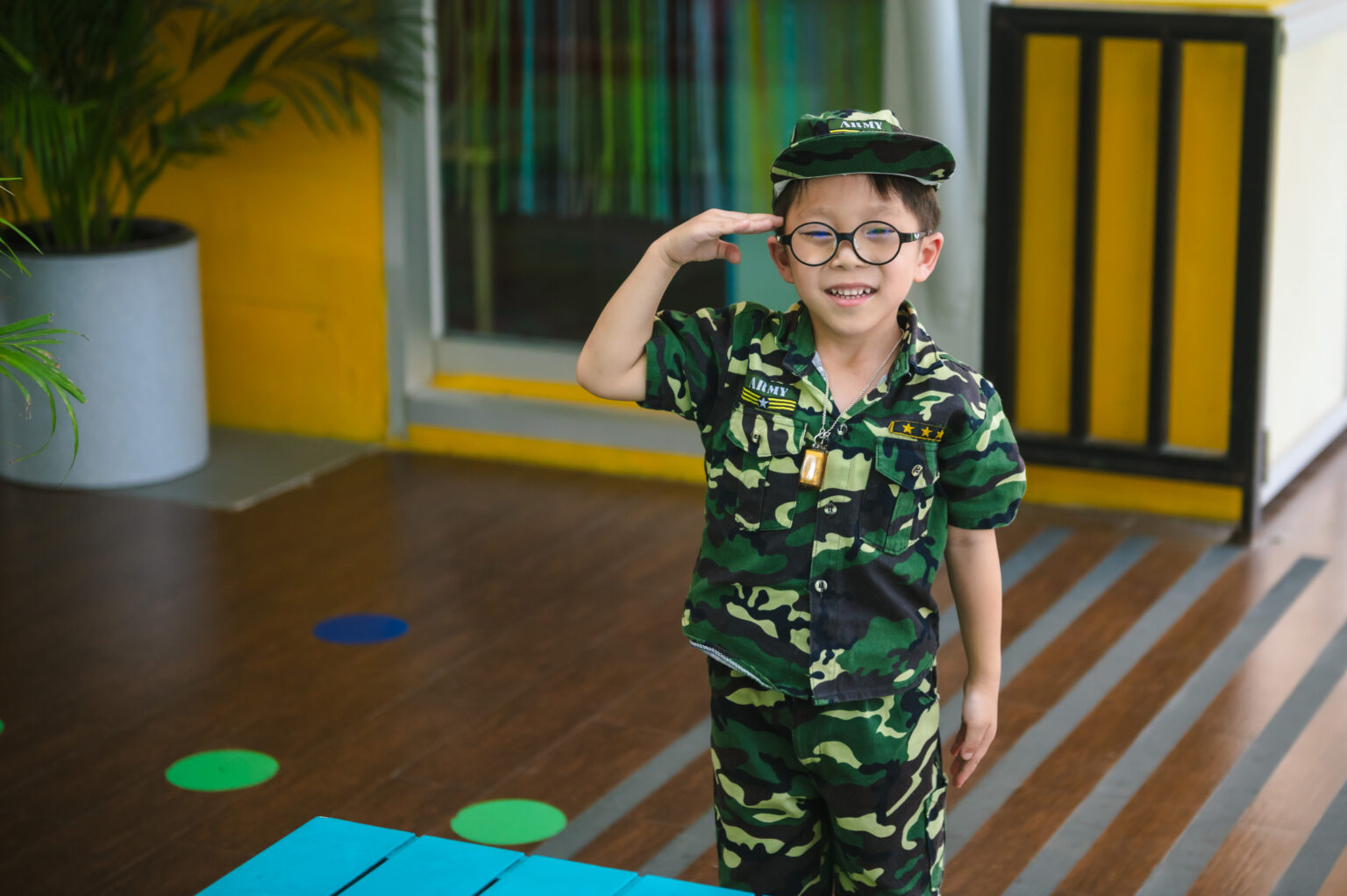 A young boy at ICSN, wearing a camouflage army uniform and glasses, smiles broadly as he salutes.