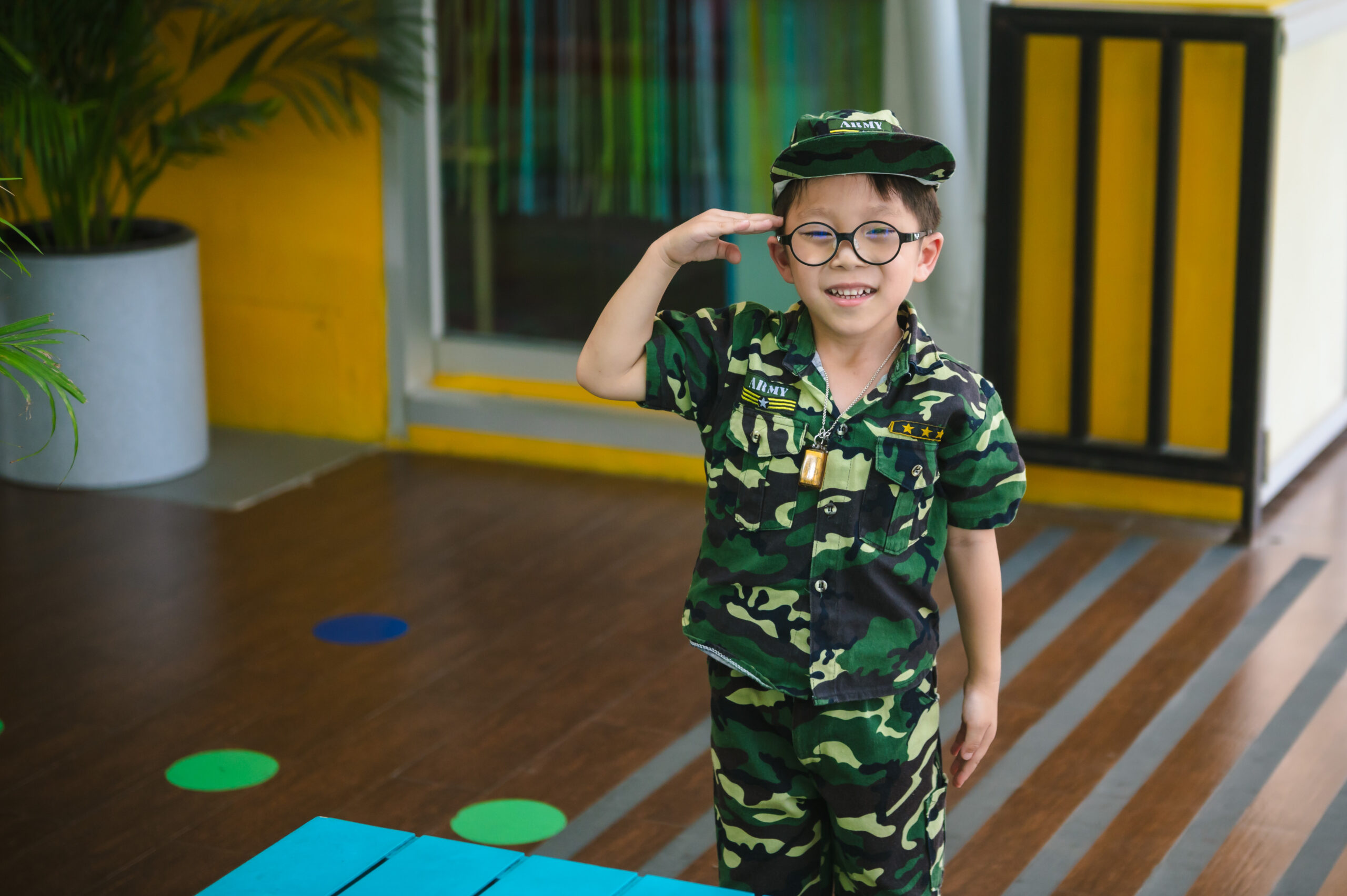 A young boy at ICSN, wearing a camouflage army uniform and glasses, smiles broadly as he salutes.