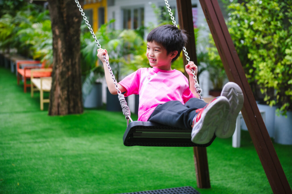 A smiling student in a pink shirt enjoys time on a swing set at the ICSN playground, surrounded by lush greenery.