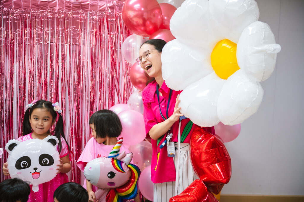 A teacher at ICSN laughs with young students while holding large colorful balloons shaped like a panda, a unicorn, and a daisy against a pink tinsel backdrop.