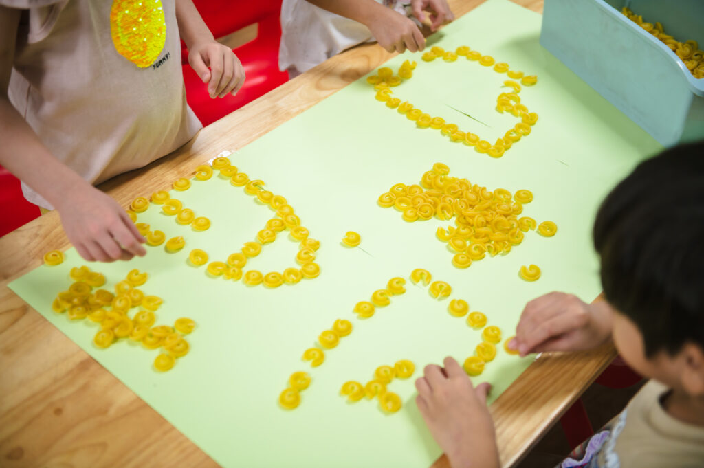Close-up of young children at ICSN using yellow pasta pieces to outline shapes on green paper during a Sensory Mathematics activity.
