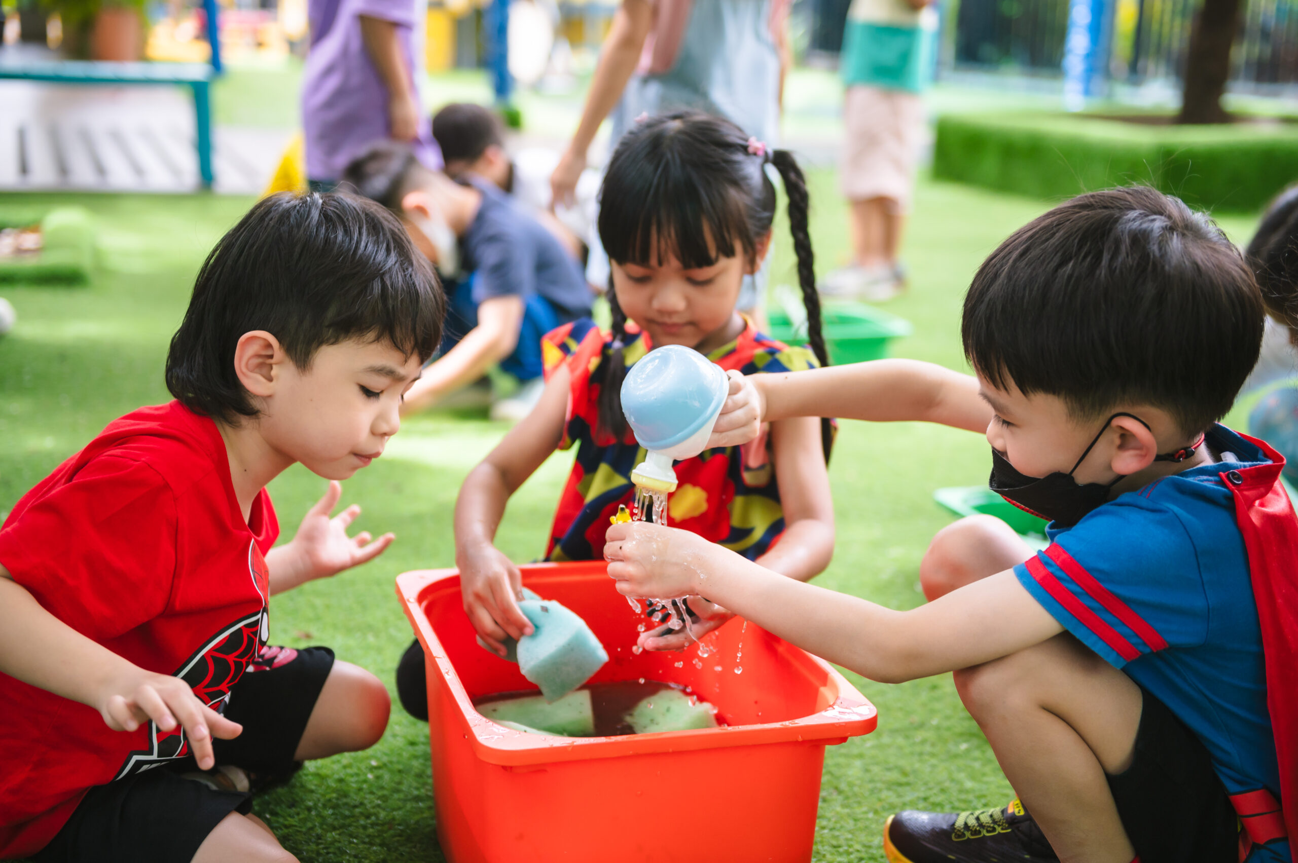 Three kindergarten students having outdoor play-based learning with water, sponges, and a red tub on a green lawn.