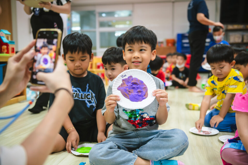 A young student proudly displays his purple clay artwork on a paper plate while a teacher takes a photo.