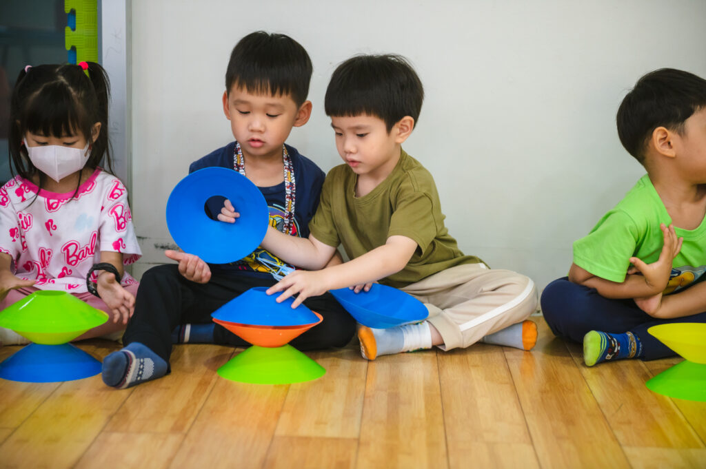 Two boys sit on a wooden floor collaborating on a construction project using blue and green plastic cones.