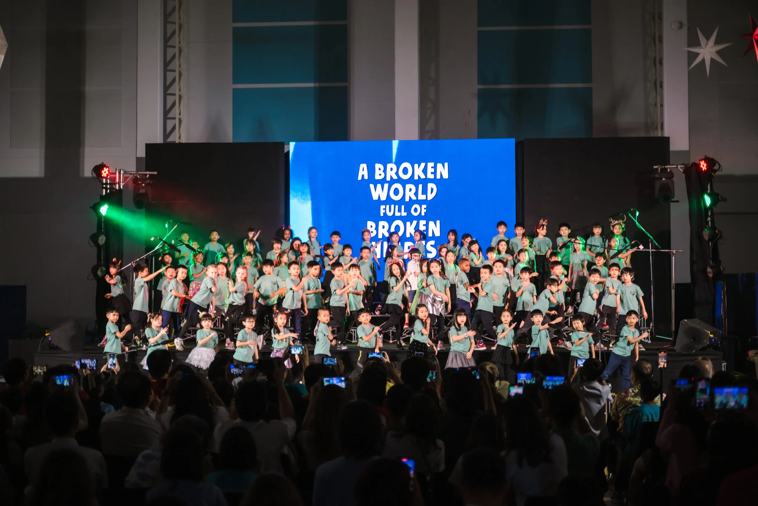 children performing a song and dance number on stage in front of a crowd of parents taking videos.