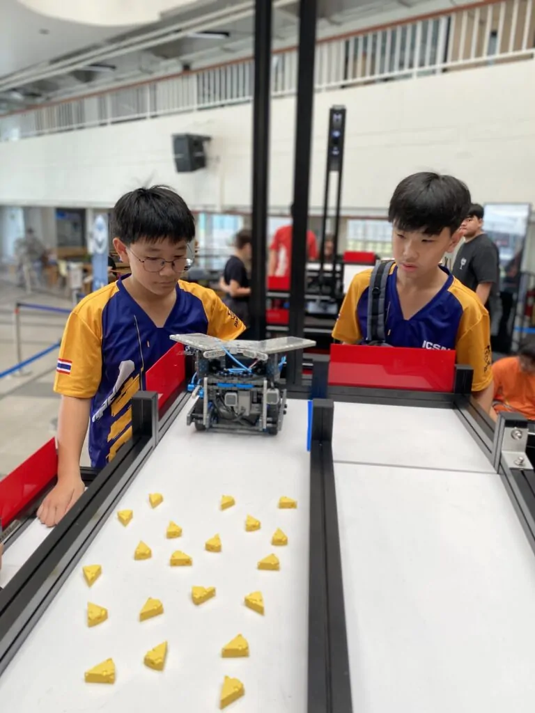 Two young boys in 'ICSN' team jerseys lean over a white competition field, intently watching a gray and blue robot. Small, yellow, cheese-wedge-shaped objects are scattered on the field in front of the robot.